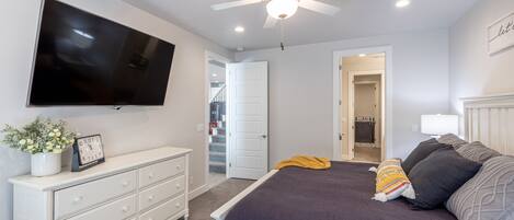 A bedroom with a bed, a dresser, a television, a ceiling fan, and a door leading to another room.