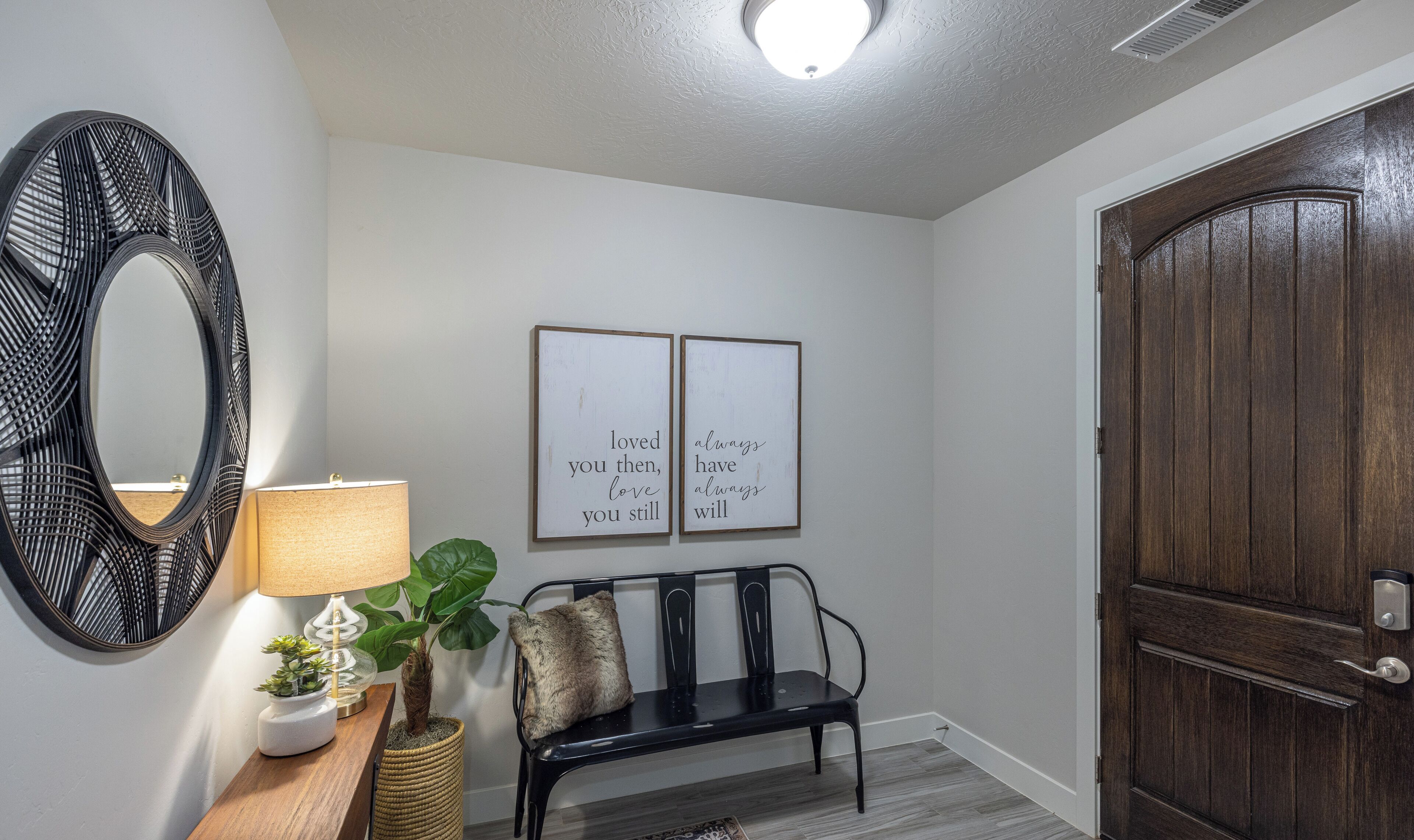 A hallway with a wooden bench, a wall-mounted mirror, a lamp, and two framed inspirational quotes.