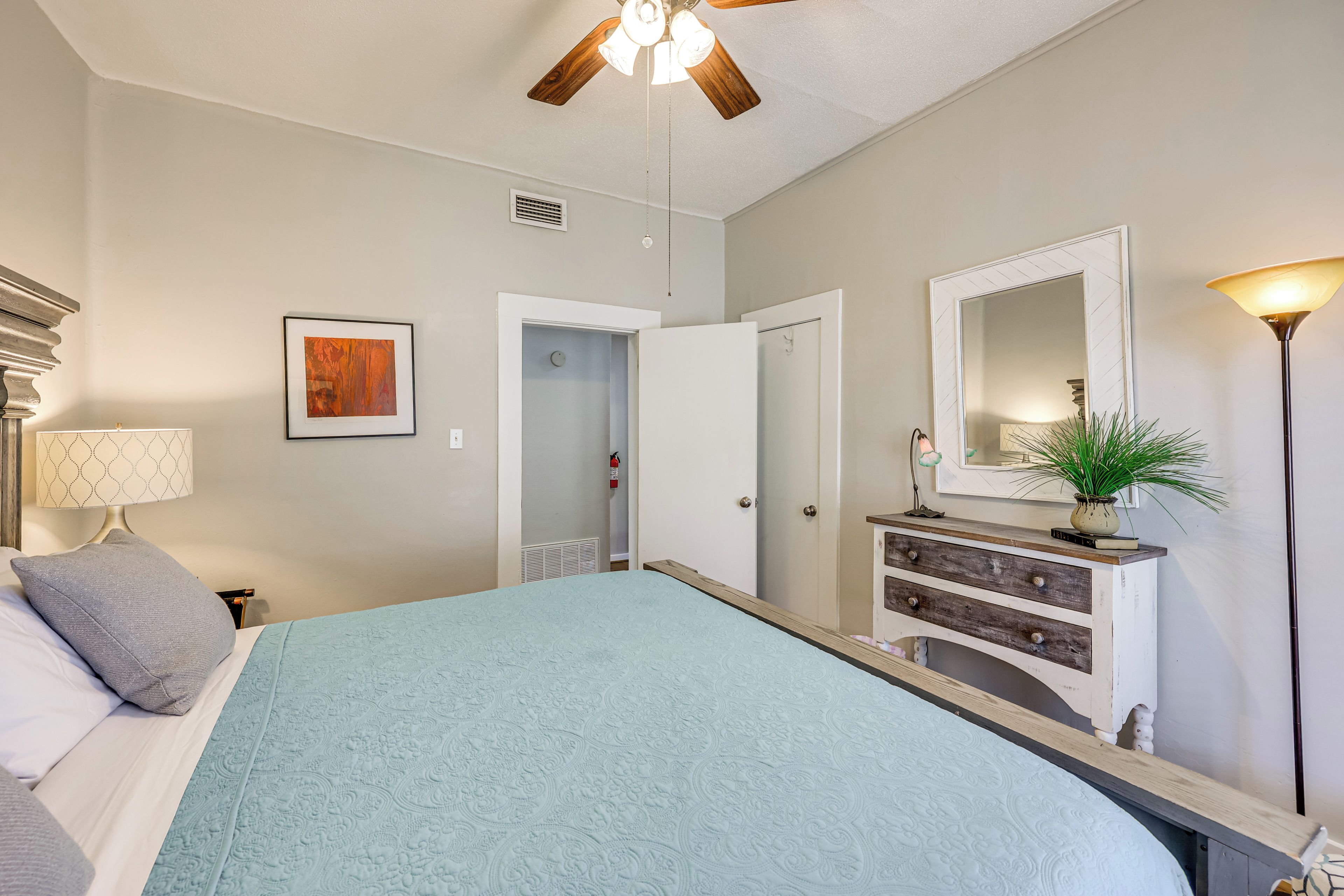Quarto com cama, cômoda, espelho, ventilador no teto e porta que dá para outro ambiente.