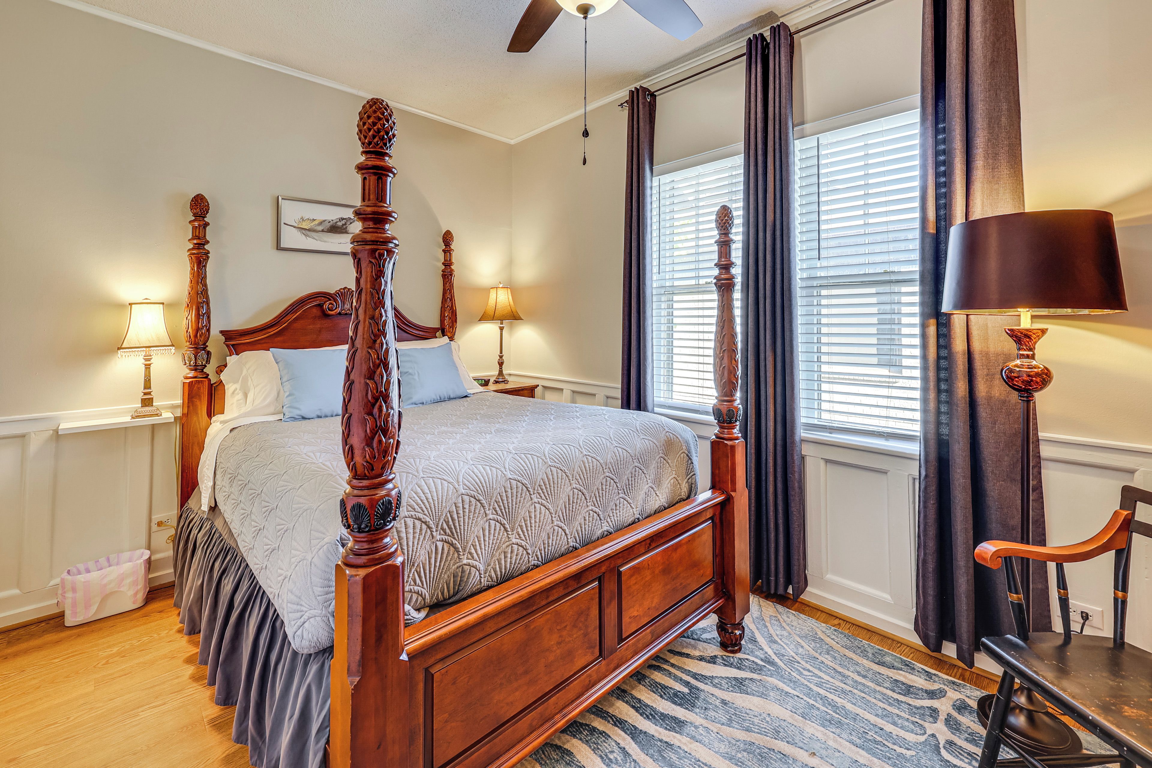 A four-poster bed with a wooden frame and a textured bedspread, a standing lamp, and a window with blinds.