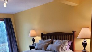 Un lit bien fait, avec une tête de lit en bois, deux tables de chevet avec des lampes, et une vue sur l’extérieur par une fenêtre ornée de rideaux.