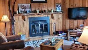 Un salon chaleureux avec un lambris en bois, une cheminée, un téléviseur fixé au mur et un coin salon confortable avec des canapés et une table basse.