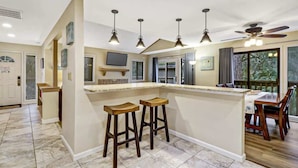 A kitchen with a central island, bar stools, pendant lights, a ceiling fan, and a dining area with a table and chairs. - 137 Beachwalk Villa Steps To The Beach (Hilton Head Island)