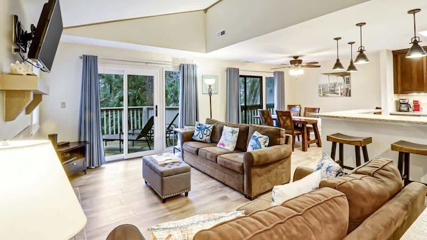 A spacious living room with a brown leather sofa, a coffee table, and a television mounted on the wall. There is a sliding glass door leading to a balcony with a view of greenery. - 137 Beachwalk Villa Steps To The Beach (Hilton Head Island)