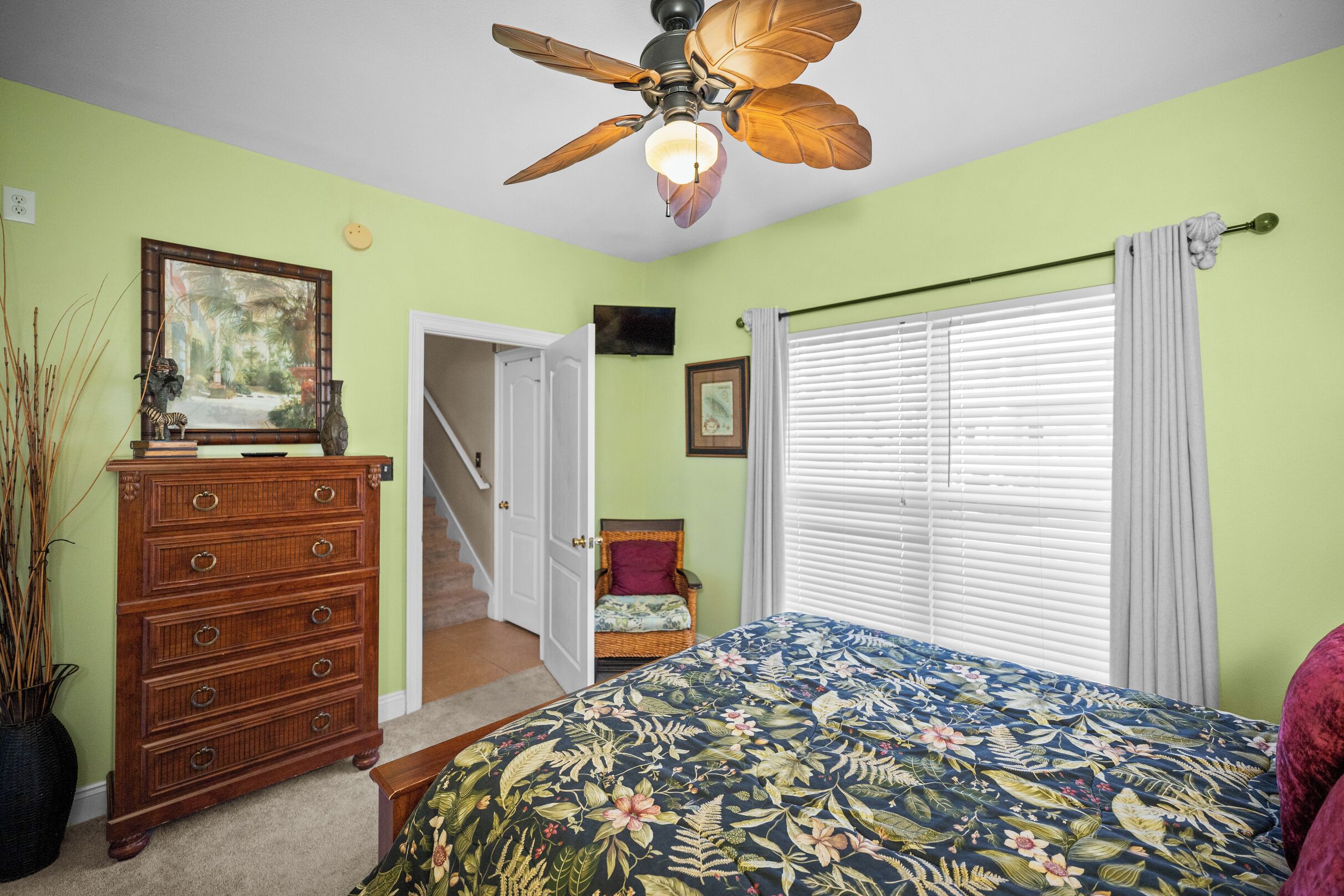 A bedroom with a bed, dresser, ceiling fan, and a staircase leading to another room.