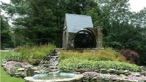 Un bâtiment rustique en pierre, doté d’une grande roue à eau, d’une petite cascade et d’un bassin circulaire avec des pierres plates permettant de le traverser à pied.