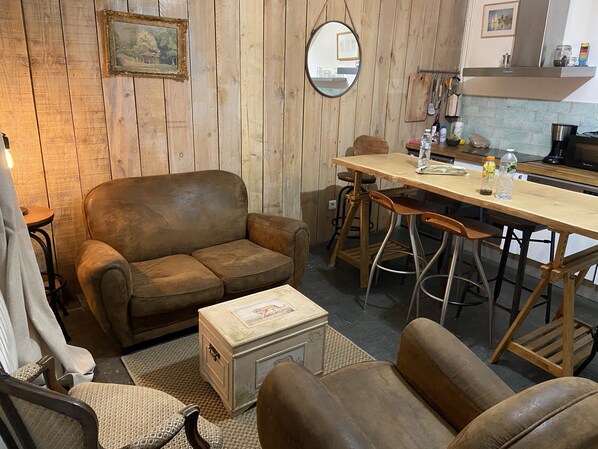 A cozy living room with a brown leather sofa, a wooden dining table with bar stools, a small wooden chest, and a mirror on the wall.
