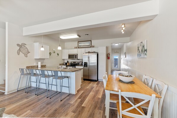 A modern kitchen with white cabinets, a central island, bar stools, a wooden dining table, and a view into the living area. - Pet-Friendly Townhome | Fenced Patio, Heated Pool & Beach Nearby (Corpus Christi)