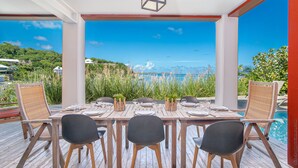 Una terraza de madera con una mesa lista para comer, con vistas panorámicas al océano y follaje.
