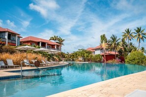 A swimming pool with lounge chairs and umbrellas, surrounded by palm trees and buildings with red roofs.