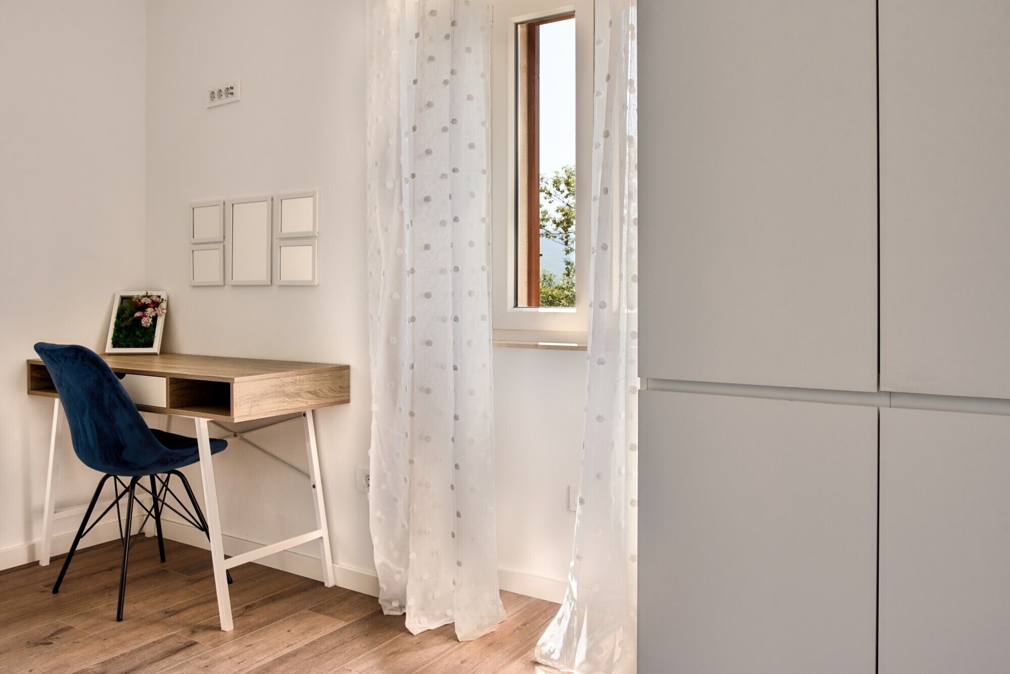 A small, bright room with a wooden desk, a blue chair, and a window with a view of trees.