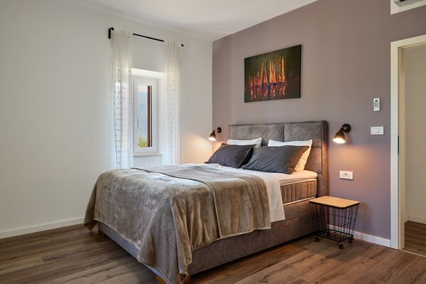 Un dormitorio con cama, mesita de noche, un cuadro y ventana con cortinas.