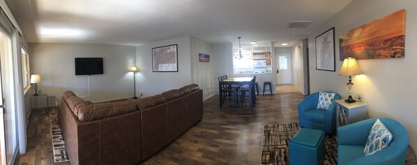 A living room with a brown sectional sofa, a blue armchair, a dining area with a table and chairs, and a kitchen in the background.