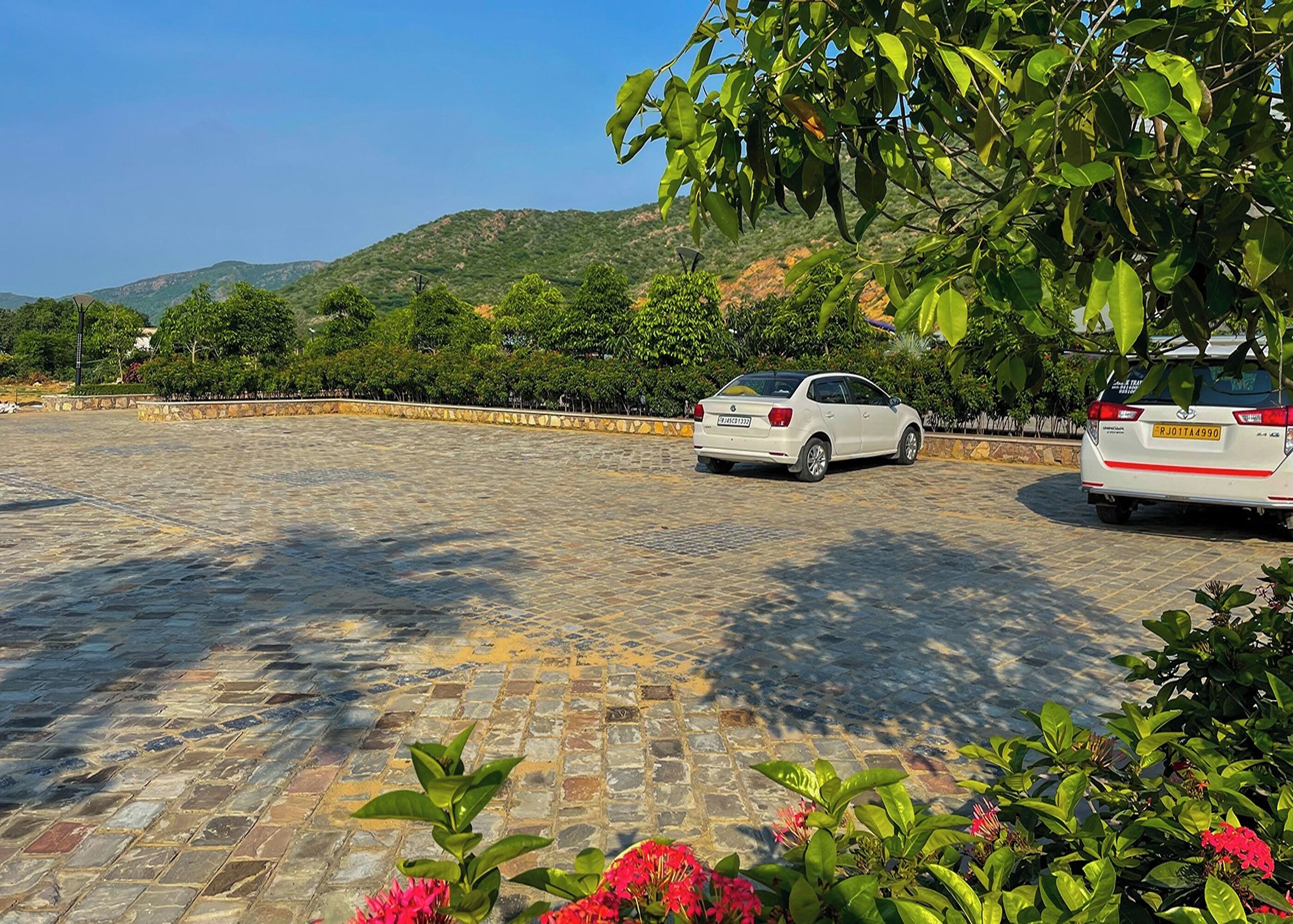 A paved parking area with two white cars parked, surrounded by greenery and hills in the background.