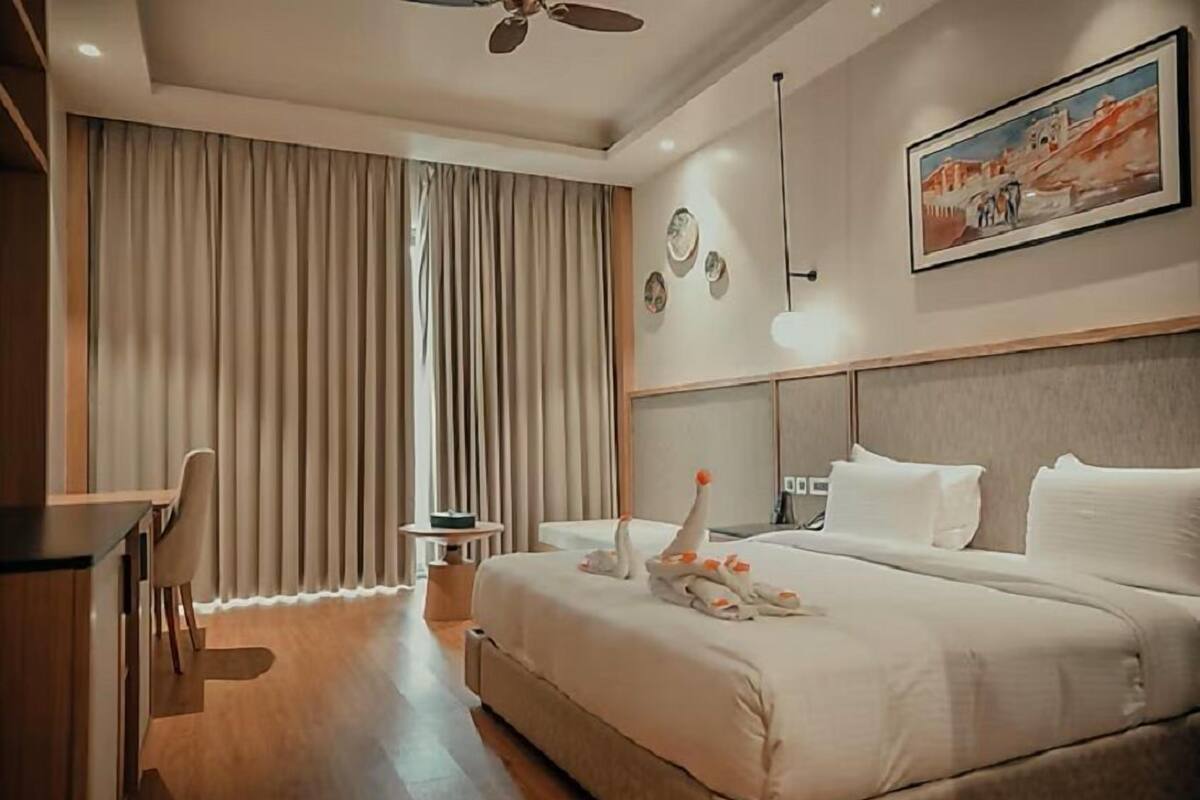 A hotel room with a bed, a desk, a chair, a small table, and a painting on the wall.