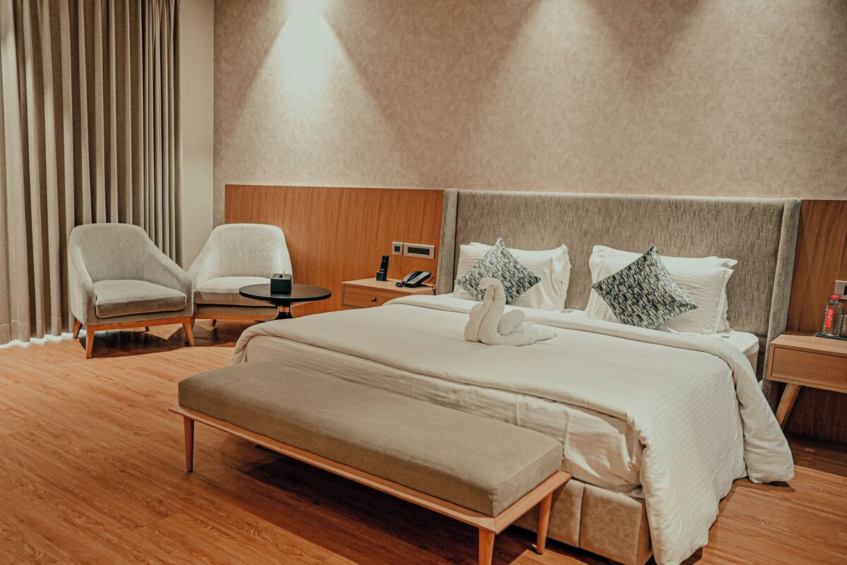 A modern hotel room with a large bed, a bedside table, a chair, a bench, and a wooden floor.