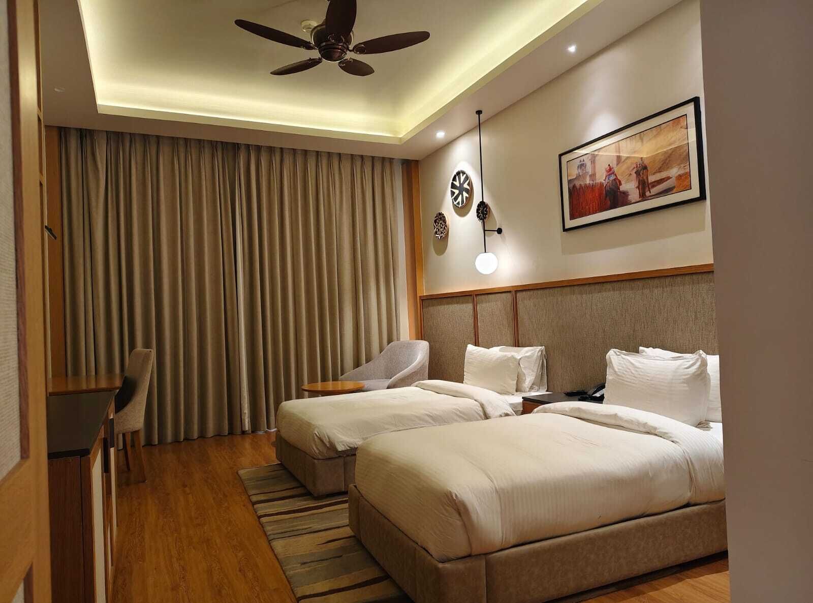 Habitación de hotel con dos camas, un escritorio, una silla, un ventilador de techo y un cuadro en la pared.