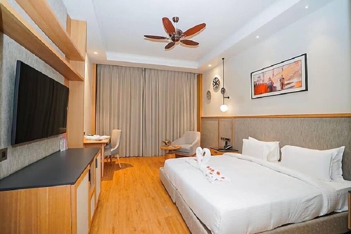 A hotel room with a bed, a desk, a chair, a TV, a ceiling fan, and a painting on the wall.