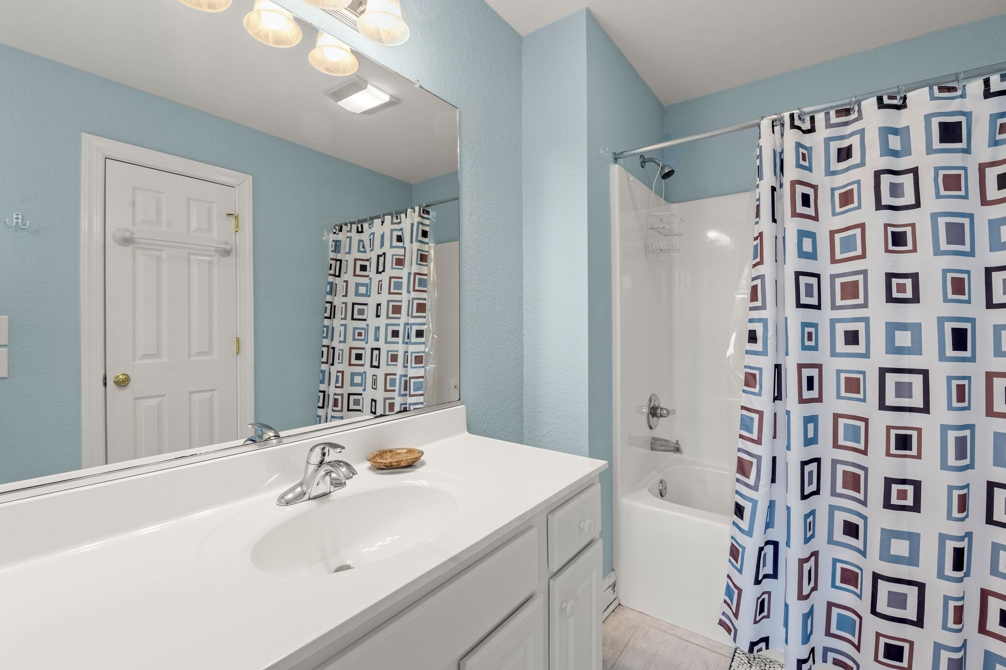 A bathroom with a white sink, a mirror, a shower with a patterned curtain, and a door.