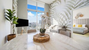 A modern living room with a large window, a flat-screen TV, a round table with a woven coaster, and a sofa with a city view.