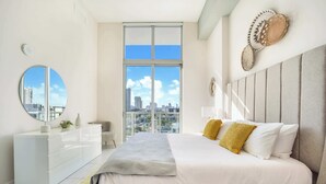 A modern bedroom with a large bed, a white dresser, a round mirror, and a city view through a large window.