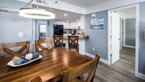 A dining area with a wooden table and chairs, a kitchen in the background, and a hallway leading to another room.