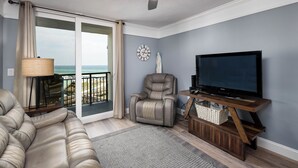A living room with a leather sofa, a recliner, a TV on a wooden stand, a clock on the wall, and a view of the ocean through a sliding door.