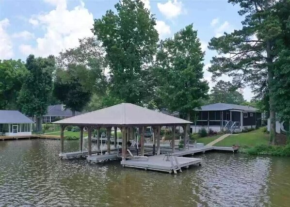 Un belvédère au bord du lac, avec une jetée qui s’avance sur l’eau, entouré d’arbres et avec une maison en arrière-plan.