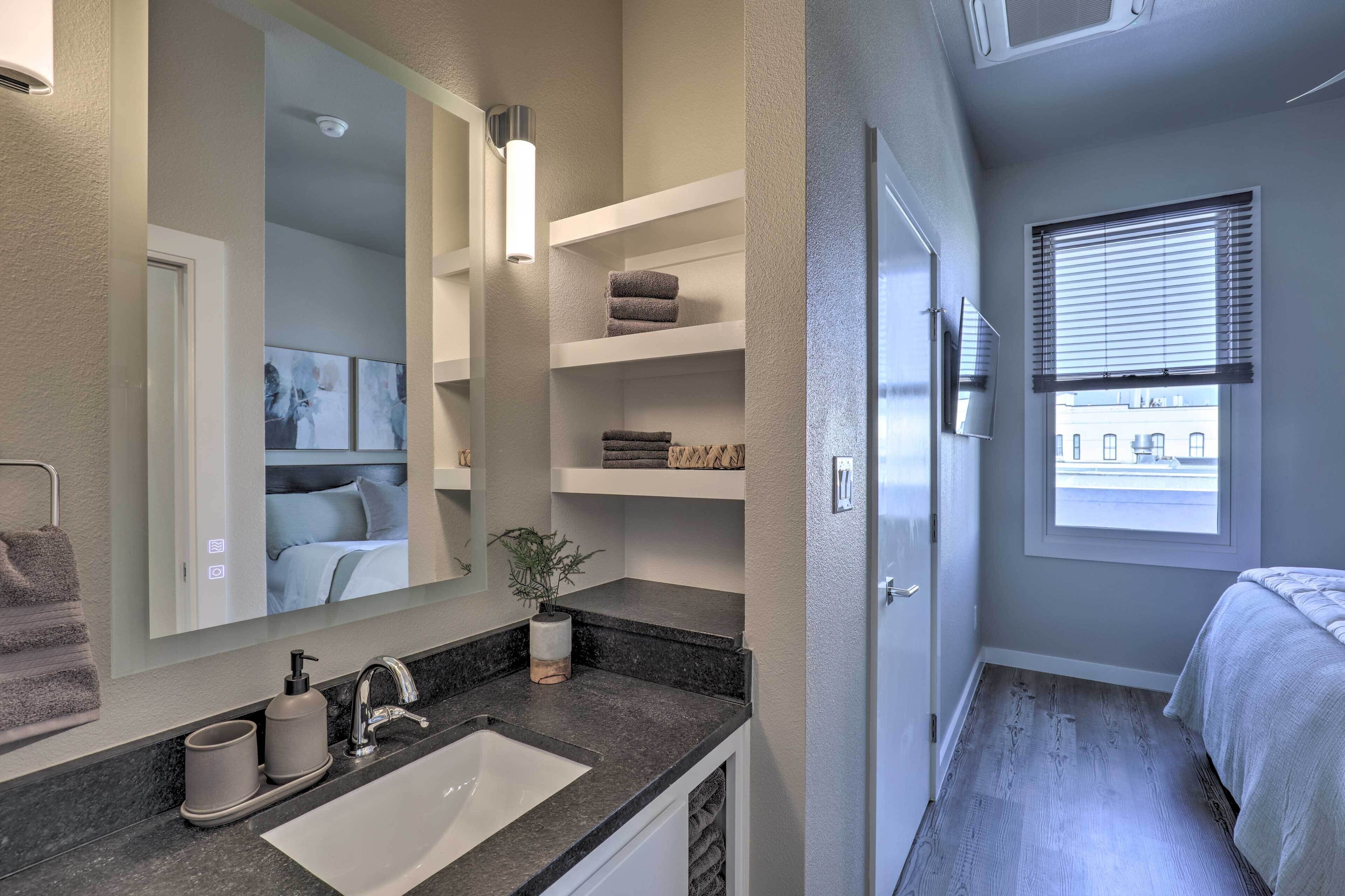 Une salle de bain moderne avec un grand miroir, un lavabo, des étagères avec des serviettes et une fenêtre avec des stores.