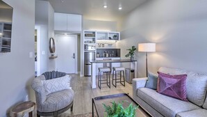 A modern living room with a grey sofa, a coffee table with plants, and a kitchen area with a bar and stools. - Centrally Located San Marcos Unit w/ Parking! (San Marcos)
