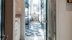 Une chambre d’hôtel moderne dotée d’une douche à parois de verre, d’une salle de bain ouverte avec pomme de douche, d’un lit recouvert d’un tapis à motifs bleus et d’une fenêtre donnant sur l’extérieur.