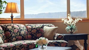 A living room with a floral sofa, a coffee table with a vase of flowers, a lamp, and a view of the mountains through a large window.