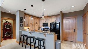 A modern kitchen with wooden cabinets, a black refrigerator, a microwave, and a sink. There are three bar stools and a trash can.