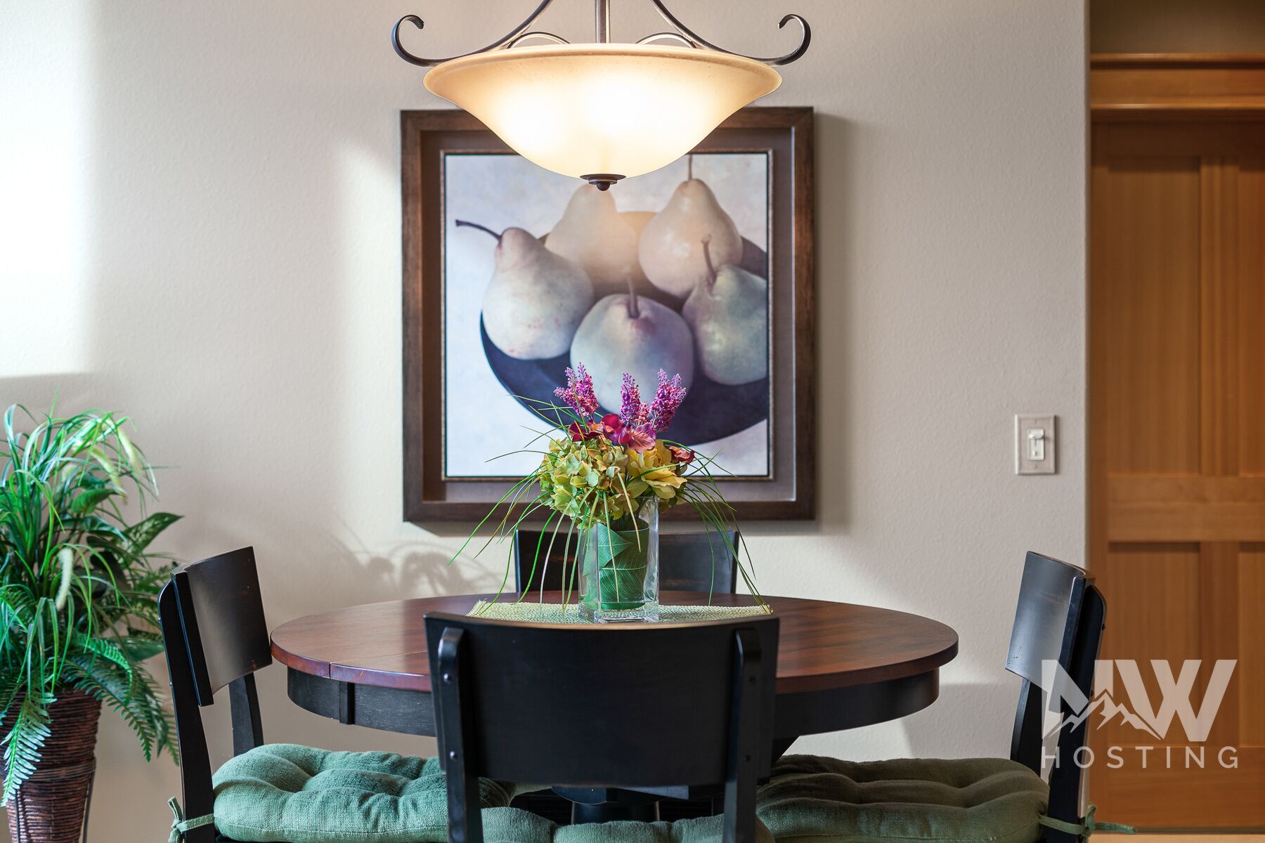 Un coin repas comprenant une table, des chaises, un vase avec des fleurs, un tableau représentant des poires accroché au mur et un luminaire au plafond.