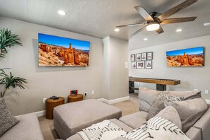 A living room with a ceiling fan, two large wall-mounted TVs displaying desert landscapes, a sofa with patterned cushions, and a small wooden side table with a remote control. - Escondido 2343 & 2355 Retreat, Connected homes with 10 Bedrooms, sleeps 40, and endless amenities (Washington)