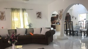 Ein modernes Wohnzimmer mit einem Glastisch, einer Couch mit Kissen, einem Ventilator und Blick in den Küchenbereich.