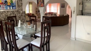 Ein Essbereich mit Glastisch und Holzstühlen, ein Wohnzimmer mit Sofa und Fernseher sowie eine Küche mit Kühlschrank.