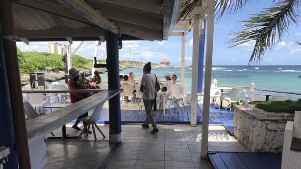 Ein überdachter Außenbereich mit Tischen und Stühlen mit Blick auf einen Strand, das Meer und ein Gebäude in der Ferne.