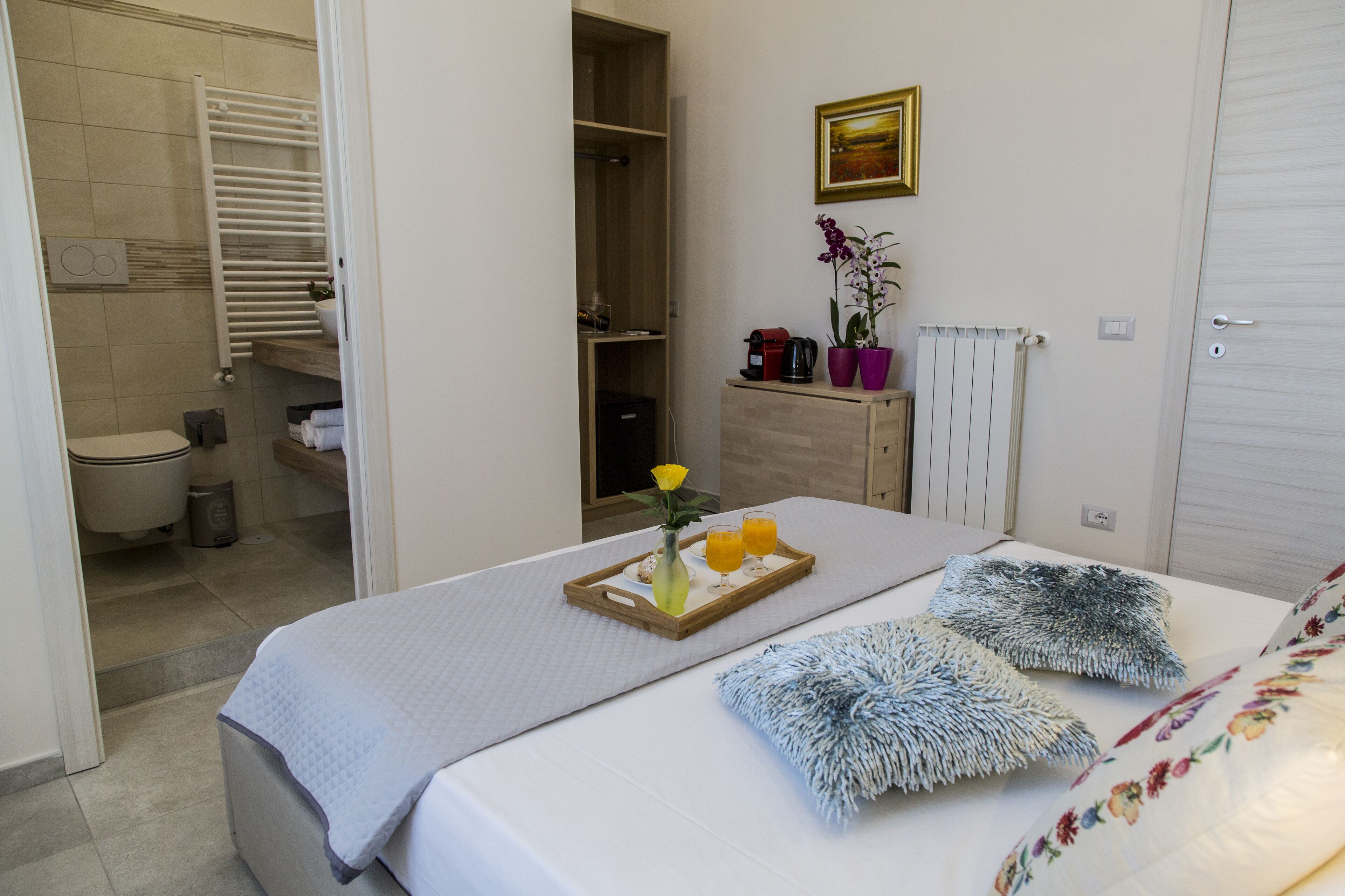 Habitación de hotel con cama, mesita de noche con un plato de jugo de naranja y un florero, radiador, armario de madera, y un baño visible a través de una puerta abierta.