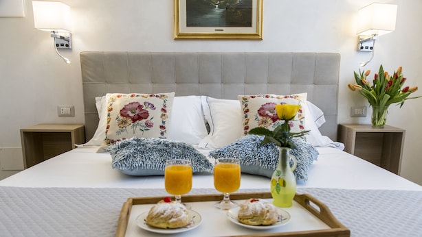 A neatly made bed with floral pillows, a bedside table with a vase of tulips, and a tray with pastries and orange juice. - Alloggio Turistico Port Inn (Civitavecchia)