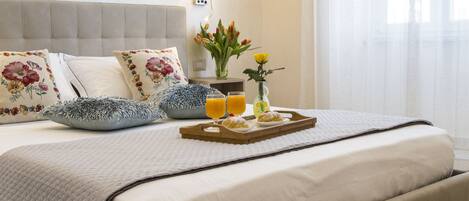A neatly made bed with floral pillows, a bedside table with a lamp and a vase of flowers, and a tray with pastries and orange juice.