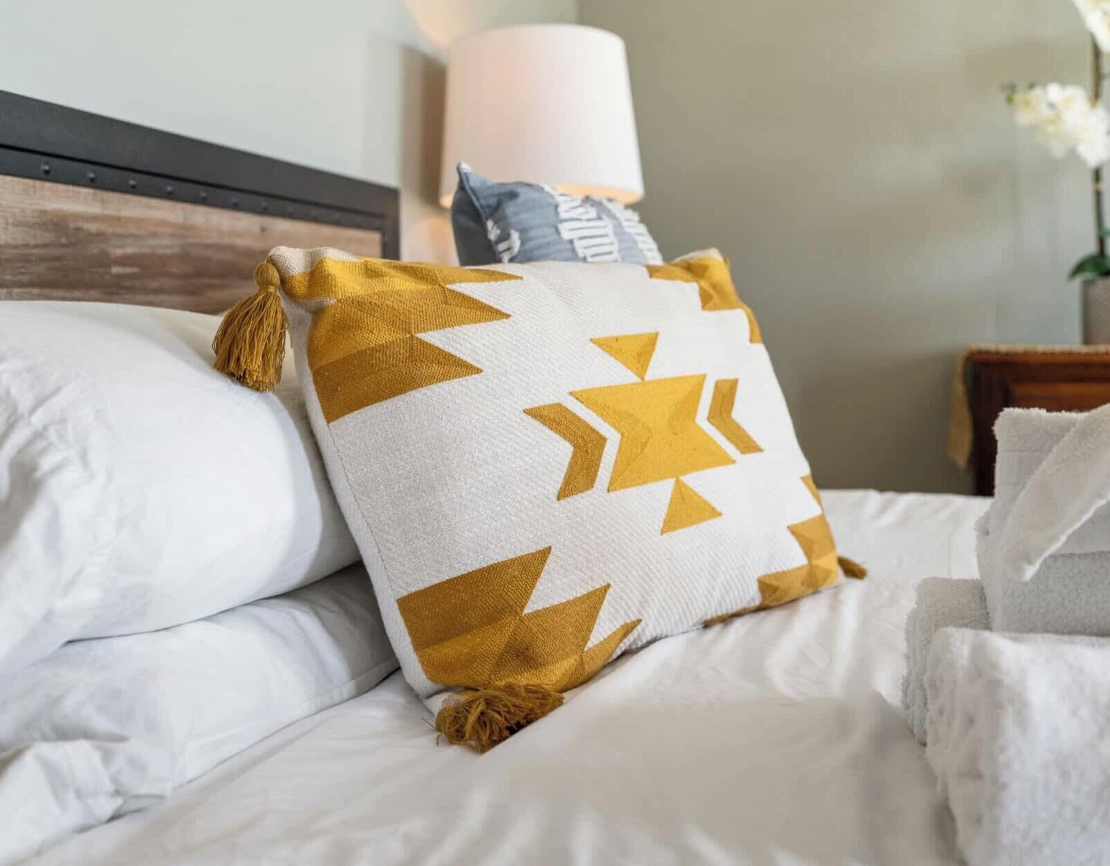 Een bed met witte beddengoed, een kussen met een geometrisch patroon en een nachtkastje met een lamp.