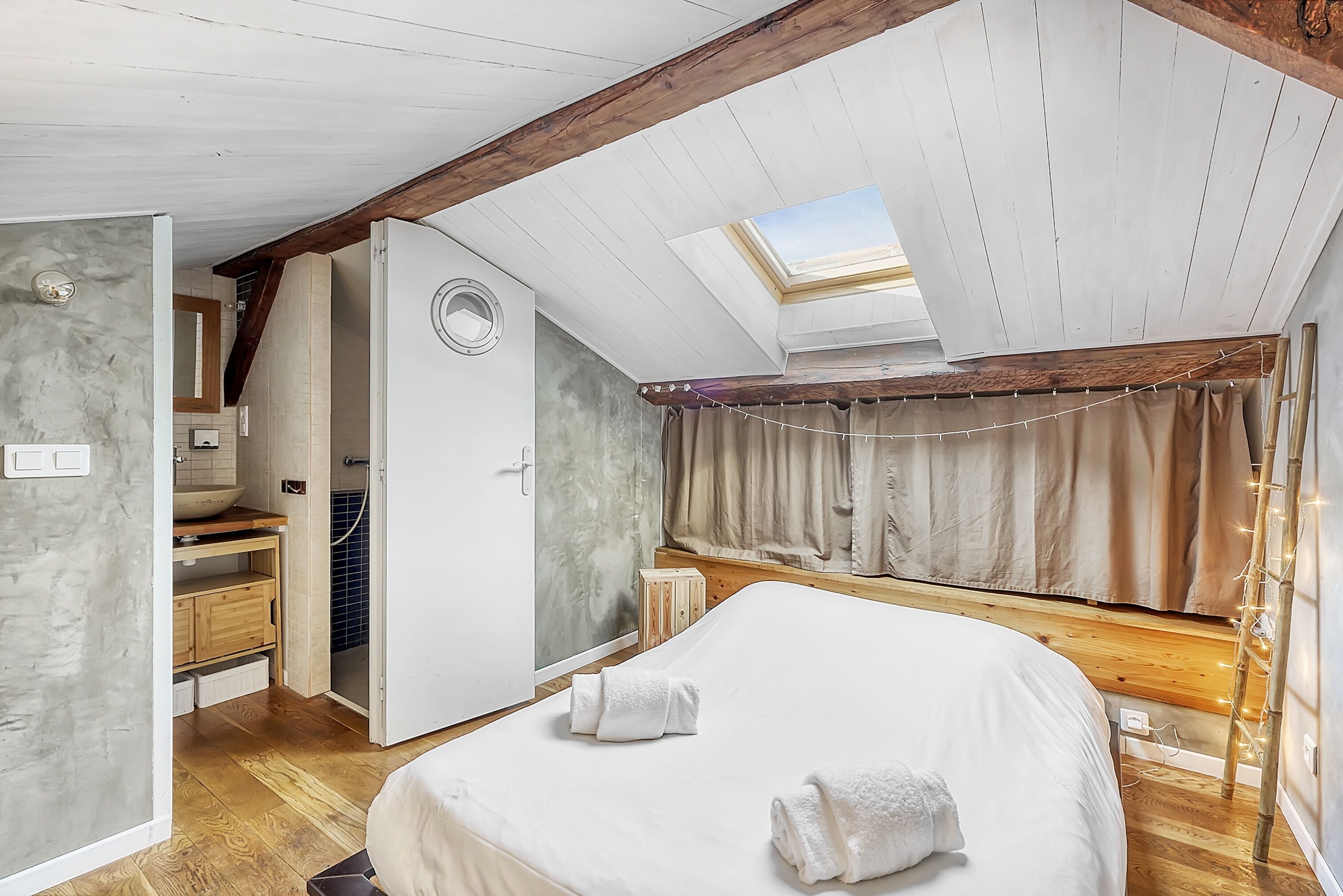 Une chambre dotée d’une lucarne, de poutres en bois, d’un lit avec des serviettes, d’un banc en bois et d’une salle de bain avec un lavabo et un miroir.