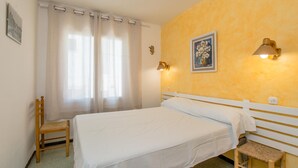 A bedroom with a bed, a chair, a small stool, a wall-mounted lamp, and a painting.