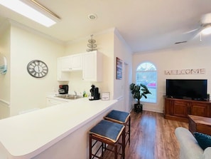 Une cuisine moderne avec des meubles blancs, un coin bar, une horloge et un téléviseur dans un espace de vie agrémenté d’une pancarte « BIENVENUE ».