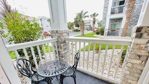 A balcony with a round table and two chairs, a white railing, and a view of a residential area with palm trees and a building with balconies. - Amazing cute & cozy condo in the center of MB (Myrtle Beach)