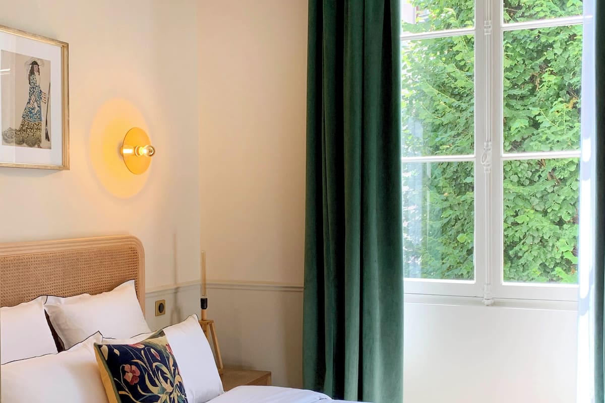 A neatly made bed with white linens and a colorful pillow, a framed artwork on the wall, and a window with green curtains.
