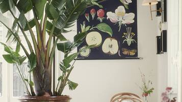 Un coin repas agrémenté d’une grande plante en pot, d’une table dressée pour deux personnes et d’un mur orné d’une illustration botanique.