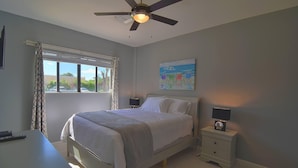 Un dormitorio con cama, mesitas de noche, ventilador de techo, ventana con cortinas y un cuadro en la pared.