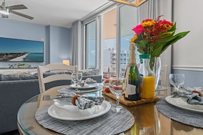 A dining area with a table set for two, featuring a bottle of champagne, a vase with flowers, and a scenic view through the window.